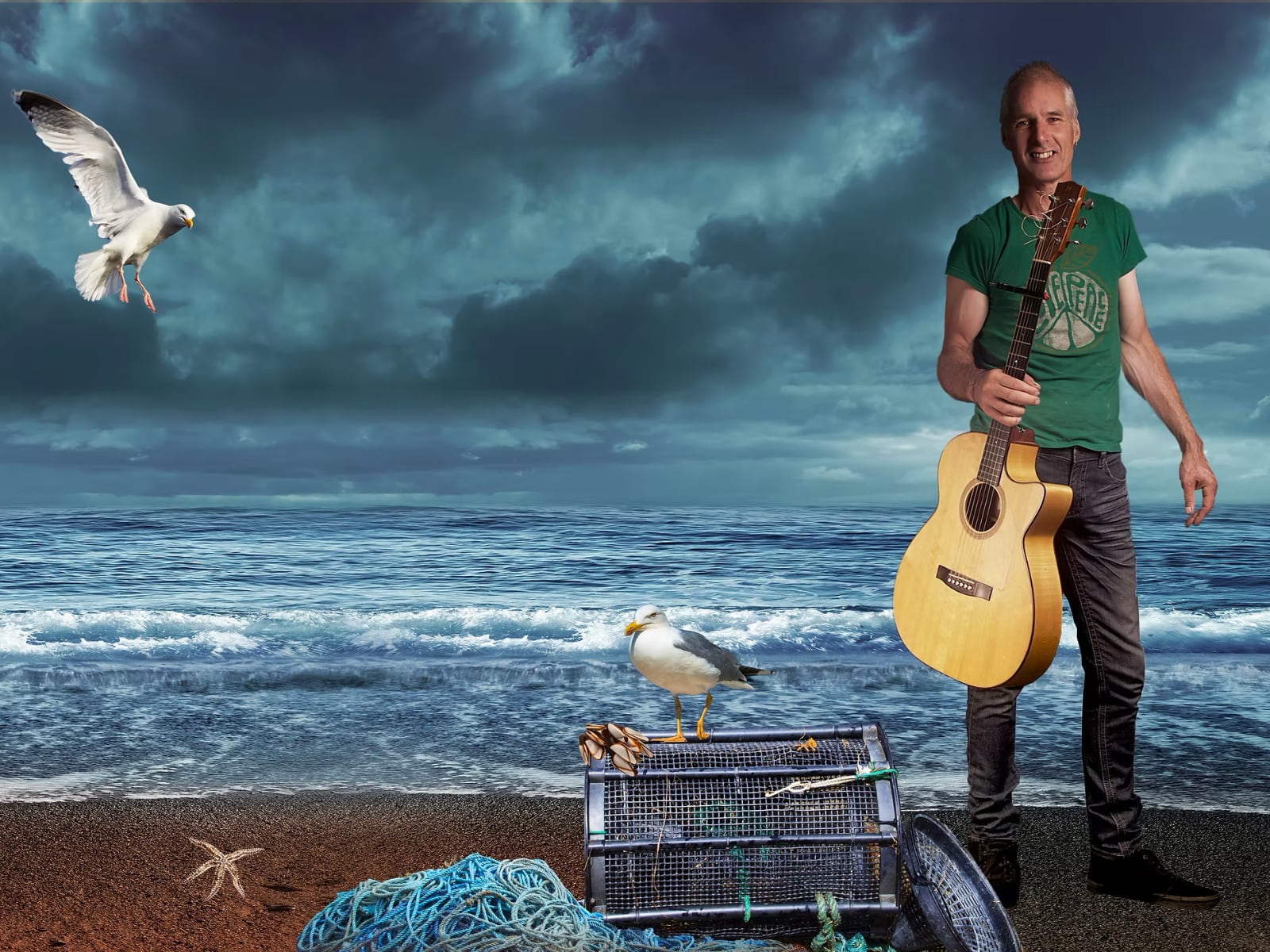 Martyn Travis standing on a beach holding his guitar with the sea behind him.