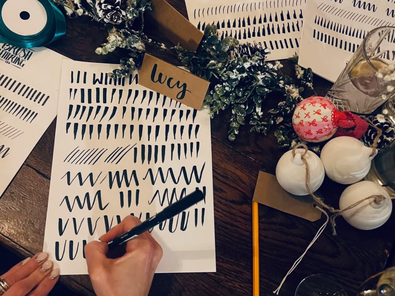 Brush lettering practice sheets being completed on a table with Christmas baubles and decorations to the side.